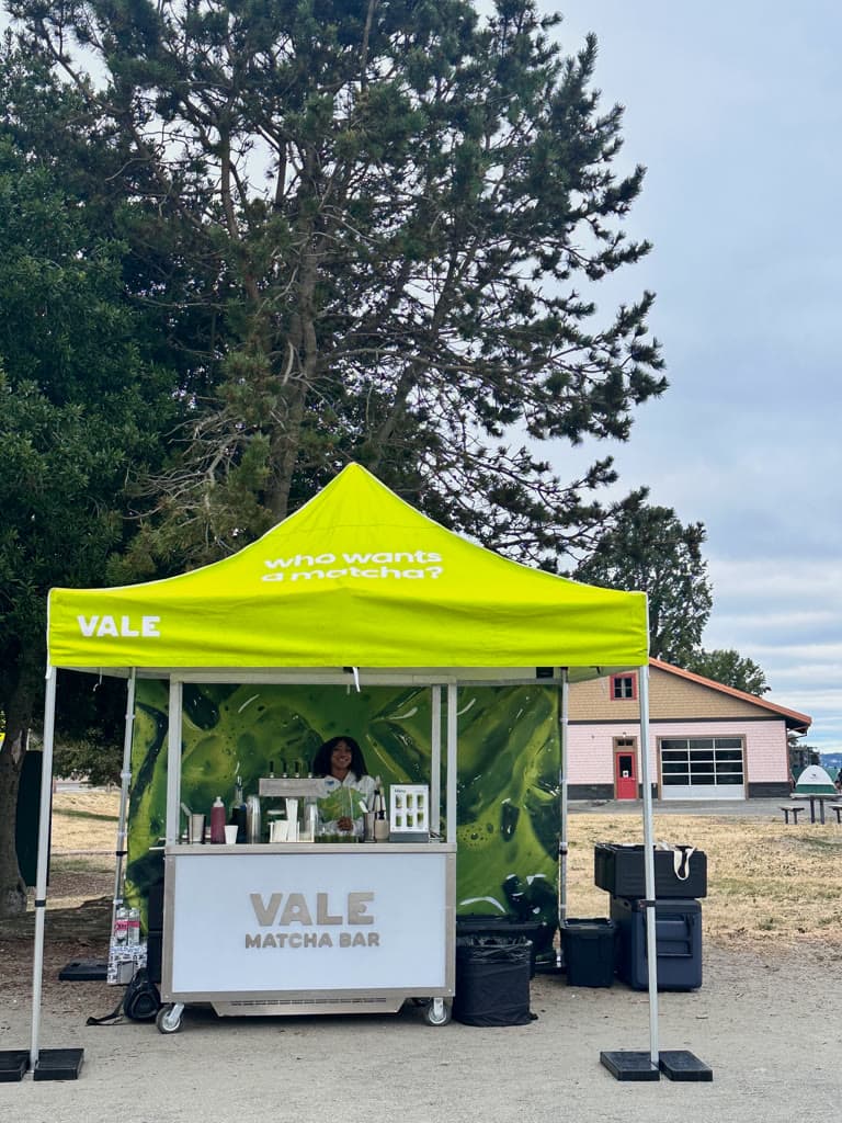 A Vale barista smiling behind the Matcha Bar cart under a lime canopy at Alki in West Seattle, with a matcha splash backdrop