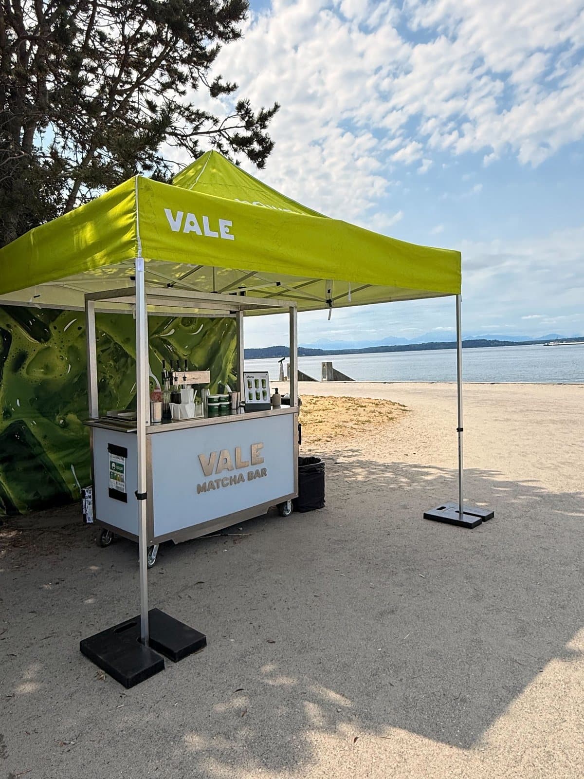 Vale Matcha Bar cart under a lime canopy on the Alki waterfront in West Seattle, with Puget Sound and mountains behind it