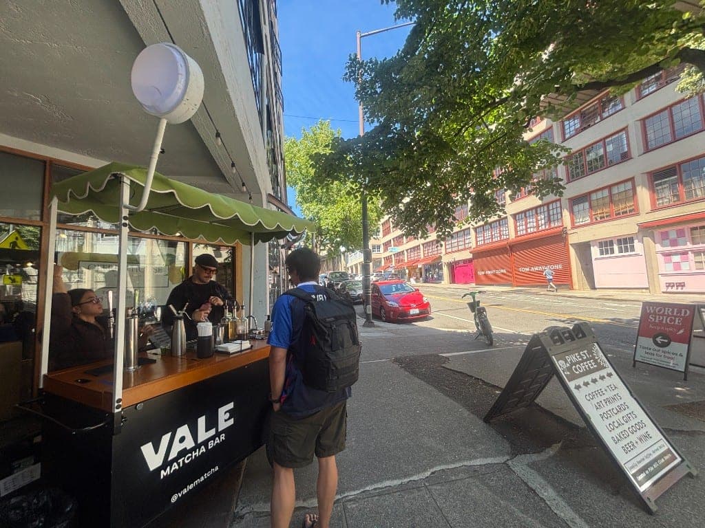 A customer ordering at the Vale Matcha Bar cart outside Citrus House near Pike Place Market, with a barista preparing matcha and the Pike St Coffee sign nearby