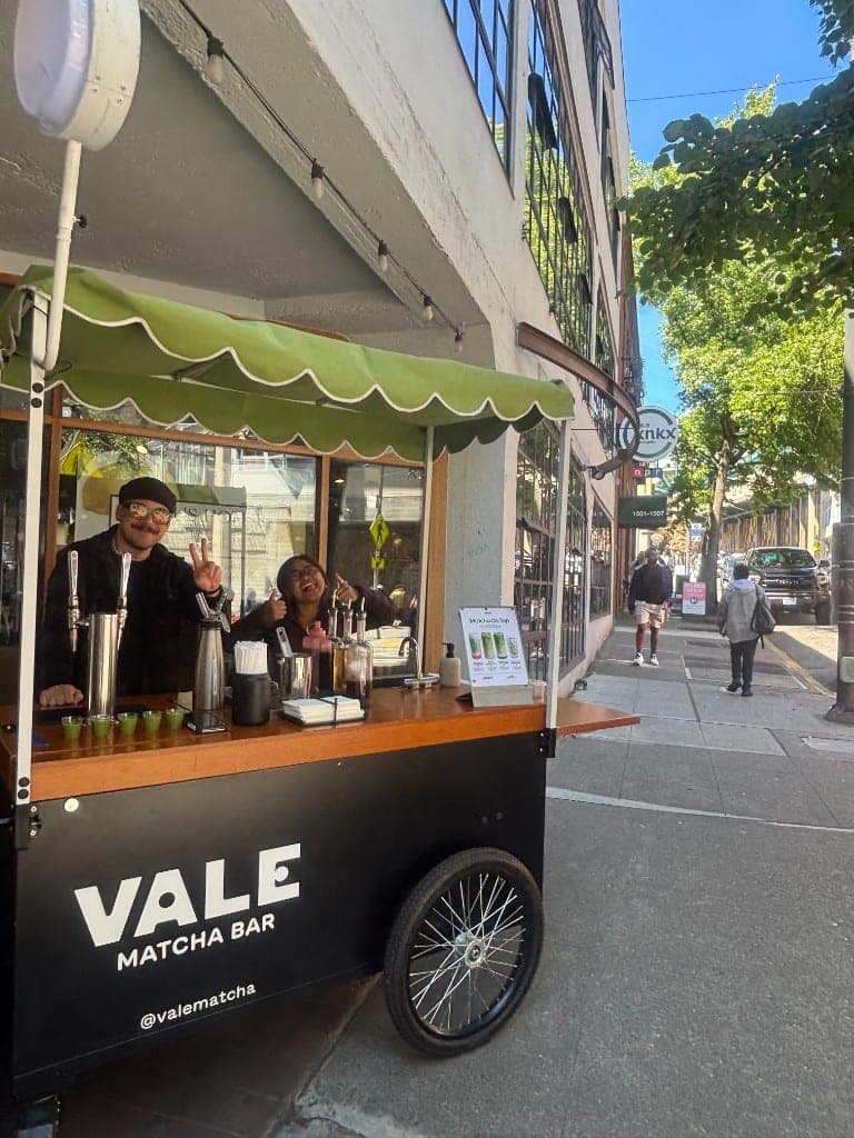 Two Vale baristas smiling behind the Vale Matcha Bar cart with its green scalloped canopy outside Citrus House on Western Ave, with matcha samples lined up on the counter
